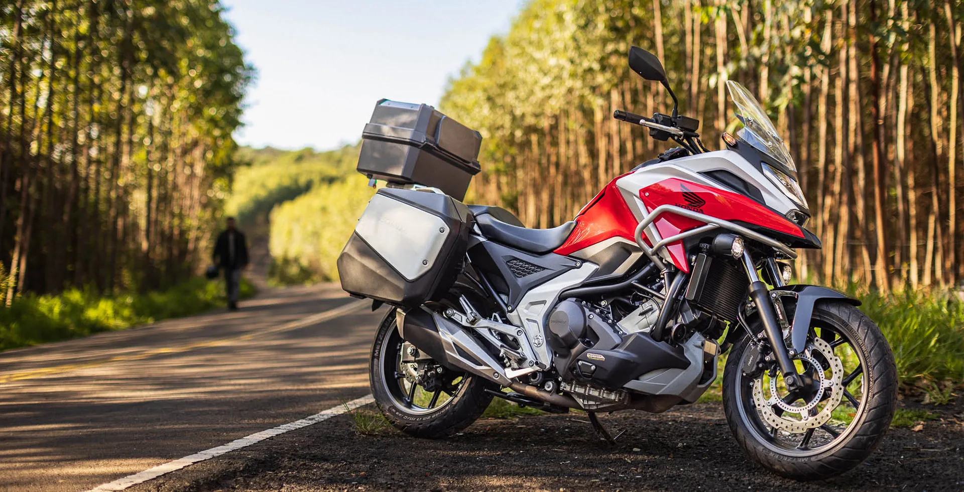 detalhe-lateral-direita-moto-honda-750x-vermelha-parada-em-estrada-arborizada-piloto-ao-fundo